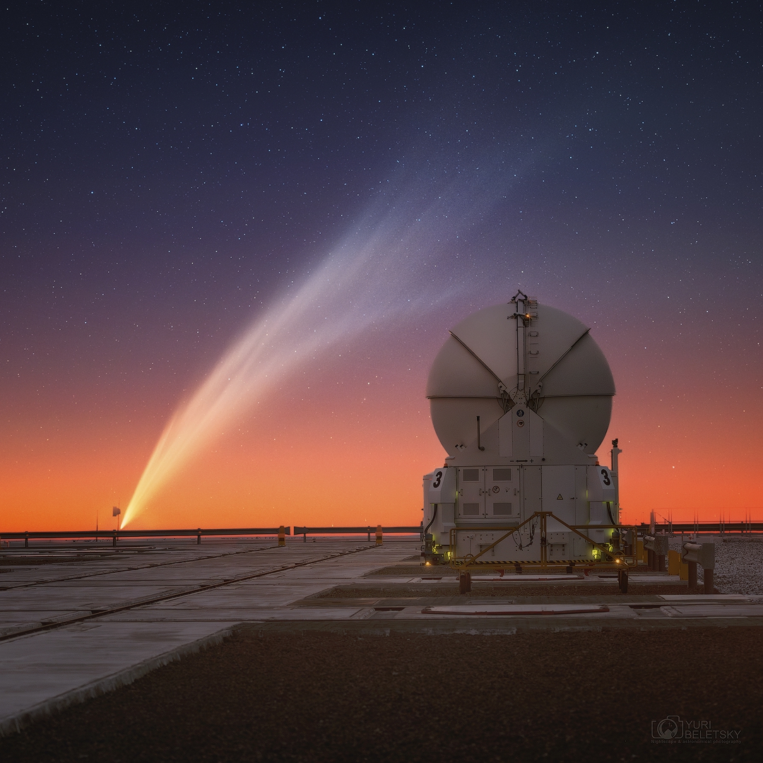 Am Horizont ragt der riesige Staubschweif über der Plattform des Paranal-Observatoriums auf. Er breitet sich diagonal über den Himmel aus, der unten orangerot leuchtet und oben in ein dunkles Blau übergeht. Rechts steht ein Hilfsteleskop mit geschlossener Kuppel.